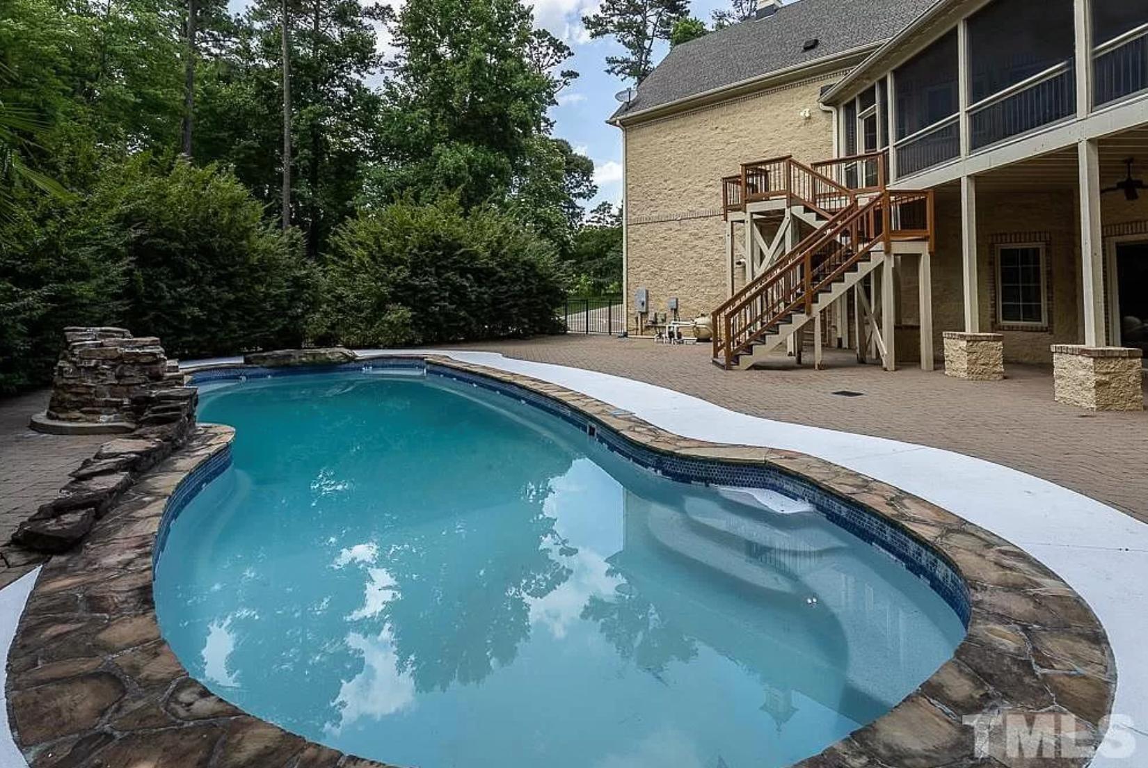 A blue pool near large, green trees and a gray, stone-faced home with a covered patio and screened-in porch